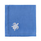 The Pink House Mustique Dinner Napkin Set features folded blue linen napkins with a white embroidered turtle in one corner, adding elegance to your luxury table linen collection.