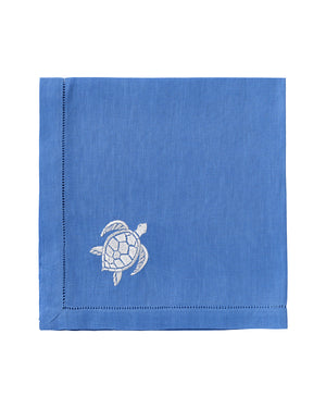The Pink House Mustique Dinner Napkin Set features folded blue linen napkins with a white embroidered turtle in one corner, adding elegance to your luxury table linen collection.
