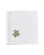 The Dinner Napkin Set by Pink House Mustique features white cloth napkins with a stitched border and a small green embroidered turtle, adding charm to your table—ideal for those seeking luxury or unique embroidered napkins.