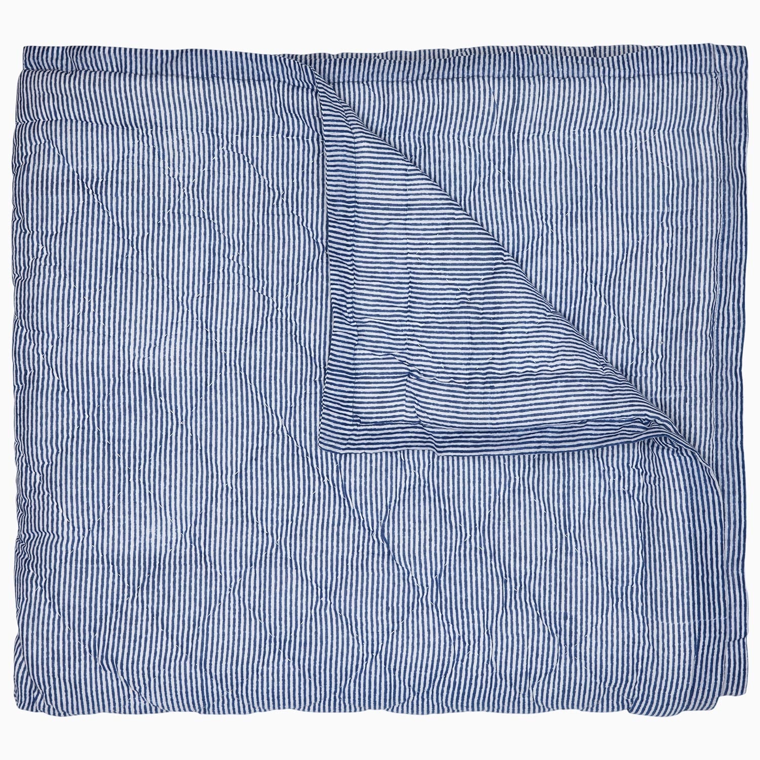 The Nandi Bedding Collection by John Robshaw is displayed partially folded back, highlighting its blue and white pinstriped design on both sides and showcasing the artisanal detail of hand-quilted craftsmanship.