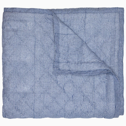 The Nandi Bedding Collection by John Robshaw is displayed partially folded back, highlighting its blue and white pinstriped design on both sides and showcasing the artisanal detail of hand-quilted craftsmanship.