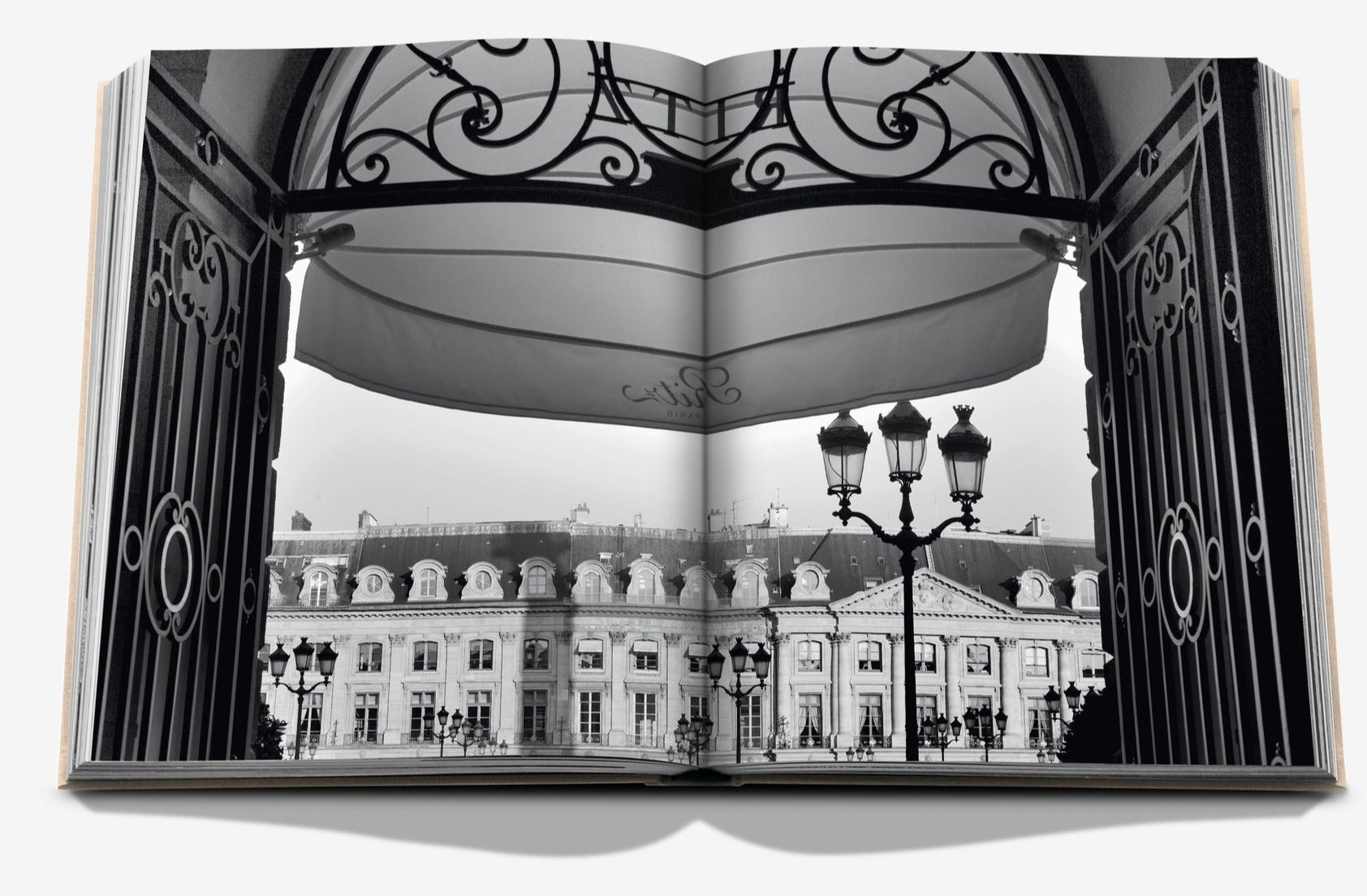 Open book displaying a black-and-white photo of the Ritz Paris, seen through ornate gates at Place Vendôme, with elegant street lamps in the foreground. The book titled "Eternally Ritz" by Assouline captures this iconic scene.