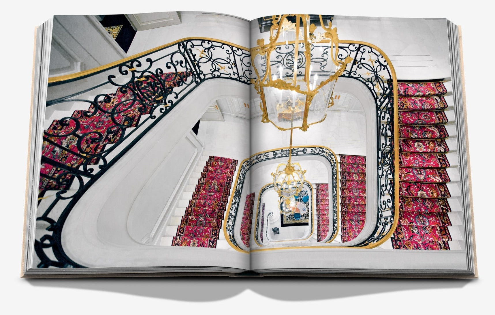 An open book displaying an ornate spiral staircase with red patterned carpet runners and intricate railing designs, viewed from above. A grand chandelier, reminiscent of the opulence found in a luxury hotel like the Ritz Paris, is also visible in the stairwell space. The book shown is "Eternally Ritz" by Assouline.