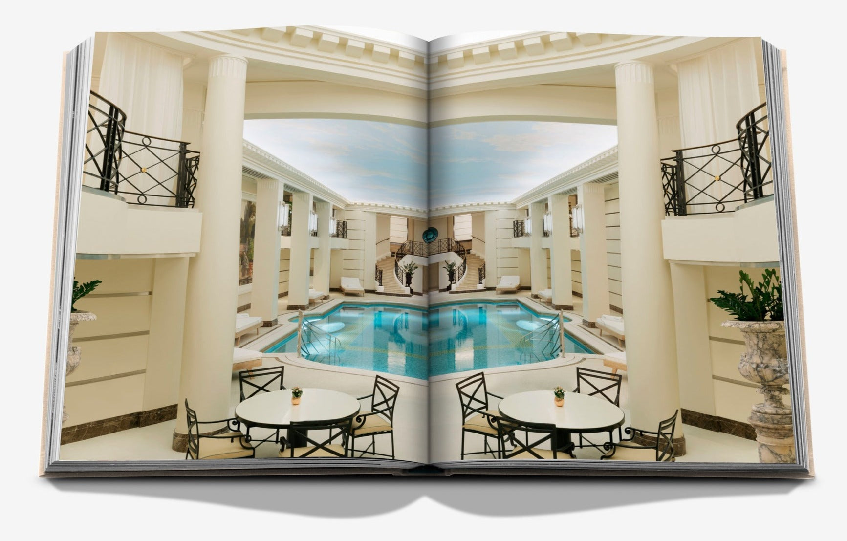 An open book displaying a photograph of an elegant indoor pool area with classical columns, decorative railings, and round tables with chairs—a scene reminiscent of the timeless beauty found in Eternally Ritz by Assouline.