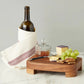 A bottle wrapped in a white towel, a glass jar with lid, knife, cheese, crackers, and green grapes are arranged on the etúHOME Classic Small Rectangle Trivet Riser displayed on a marble surface.