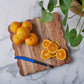 The etúHOME Scalloped Serving Board, Medium Square, displays whole and sliced oranges with a blue-handled knife next to a green potted plant on a marble surface.