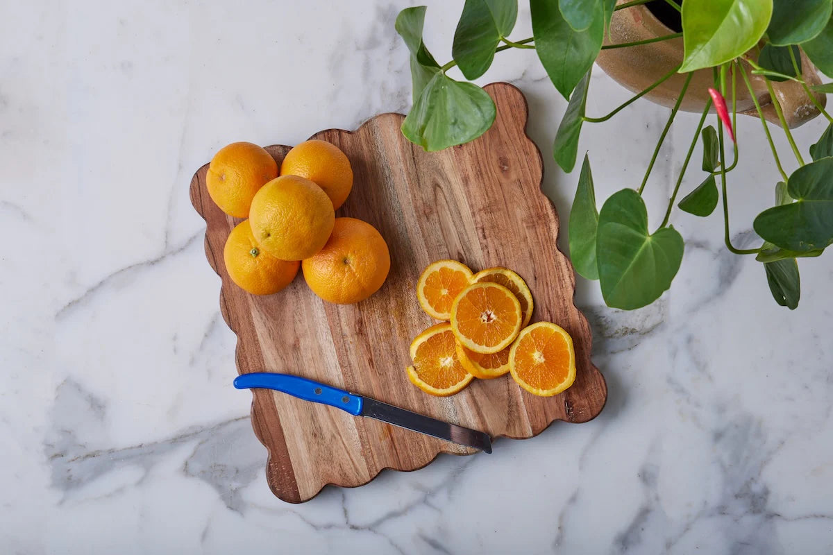 The etúHOME Scalloped Serving Board, Medium Square, displays whole and sliced oranges with a blue-handled knife next to a green potted plant on a marble surface.