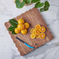 A cluster of whole and sliced oranges, green leaves, and a blue-handled knife rest on the large square Scalloped Serving Board by etúHOME, made of Acacia wood and set atop a marble surface.