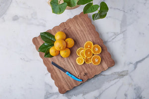 A cluster of whole and sliced oranges, green leaves, and a blue-handled knife rest on the large square Scalloped Serving Board by etúHOME, made of Acacia wood and set atop a marble surface.