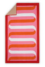 The Matouk Ribbon Beach Towel features a red border, pink background, and wavy orange and white geometric pattern. One corner folds over to reveal its soft tan cotton terry underside.