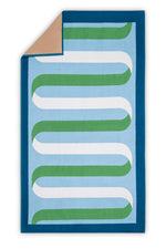The Matouk Ribbon Beach Towel features a rectangular design with a blue border, light blue background, green and white wavy lines, and a tan cotton terry underside visible at the top left corner.