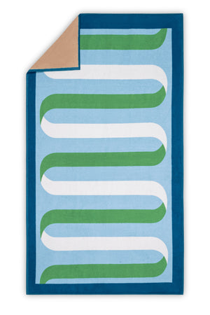 The Matouk Ribbon Beach Towel features a rectangular design with a blue border, light blue background, green and white wavy lines, and a tan cotton terry underside visible at the top left corner.