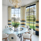 A modern dining area featuring a round glass table, white chairs, geometric chandelier, expansive windows, and gradient curtains; a potted orchid serves as the centerpiece—an inviting space where classic-meets-modern sensibilities reflect bespoke interiors inspired by Stewart Manger and beautifully showcased in Romancing the Home by Rizzoli.