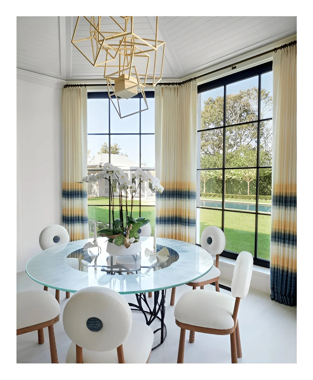 A modern dining area featuring a round glass table, white chairs, geometric chandelier, expansive windows, and gradient curtains; a potted orchid serves as the centerpiece—an inviting space where classic-meets-modern sensibilities reflect bespoke interiors inspired by Stewart Manger and beautifully showcased in Romancing the Home by Rizzoli.