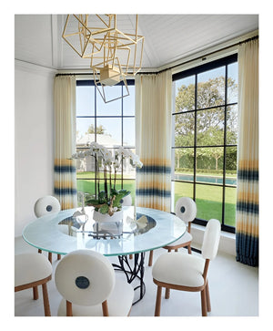 A modern dining area featuring a round glass table, white chairs, geometric chandelier, expansive windows, and gradient curtains; a potted orchid serves as the centerpiece—an inviting space where classic-meets-modern sensibilities reflect bespoke interiors inspired by Stewart Manger and beautifully showcased in Romancing the Home by Rizzoli.