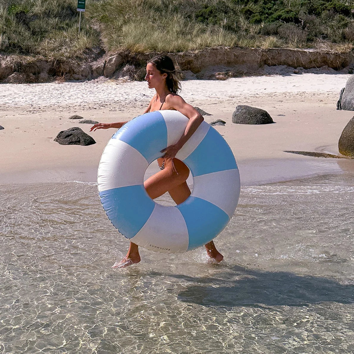 A woman in a swimsuit stands in shallow water with the Sunnylife Poolside Tube Float Blue around her waist, capturing the carefree Australian summer vibe. Sandy shore and grassy dunes frame this classic Sydney beaches scene.
