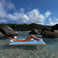 Relax in true Aussie summer style on the Sunnylife Luxe Lilo Blue Stripe inflatable, floating in clear shallow water by rocky hills beneath a partly cloudy sky.