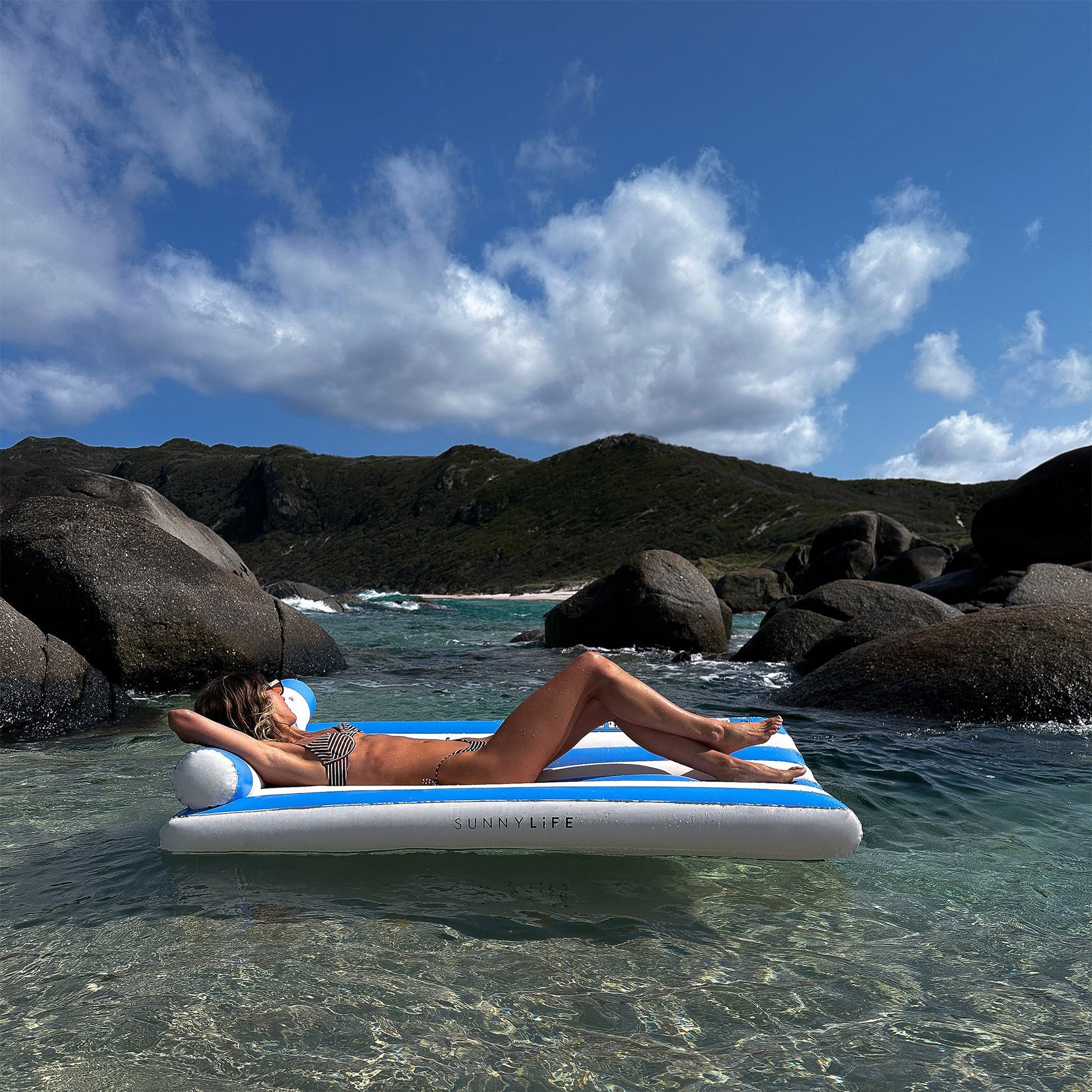 Relax in true Aussie summer style on the Sunnylife Luxe Lilo Blue Stripe inflatable, floating in clear shallow water by rocky hills beneath a partly cloudy sky.