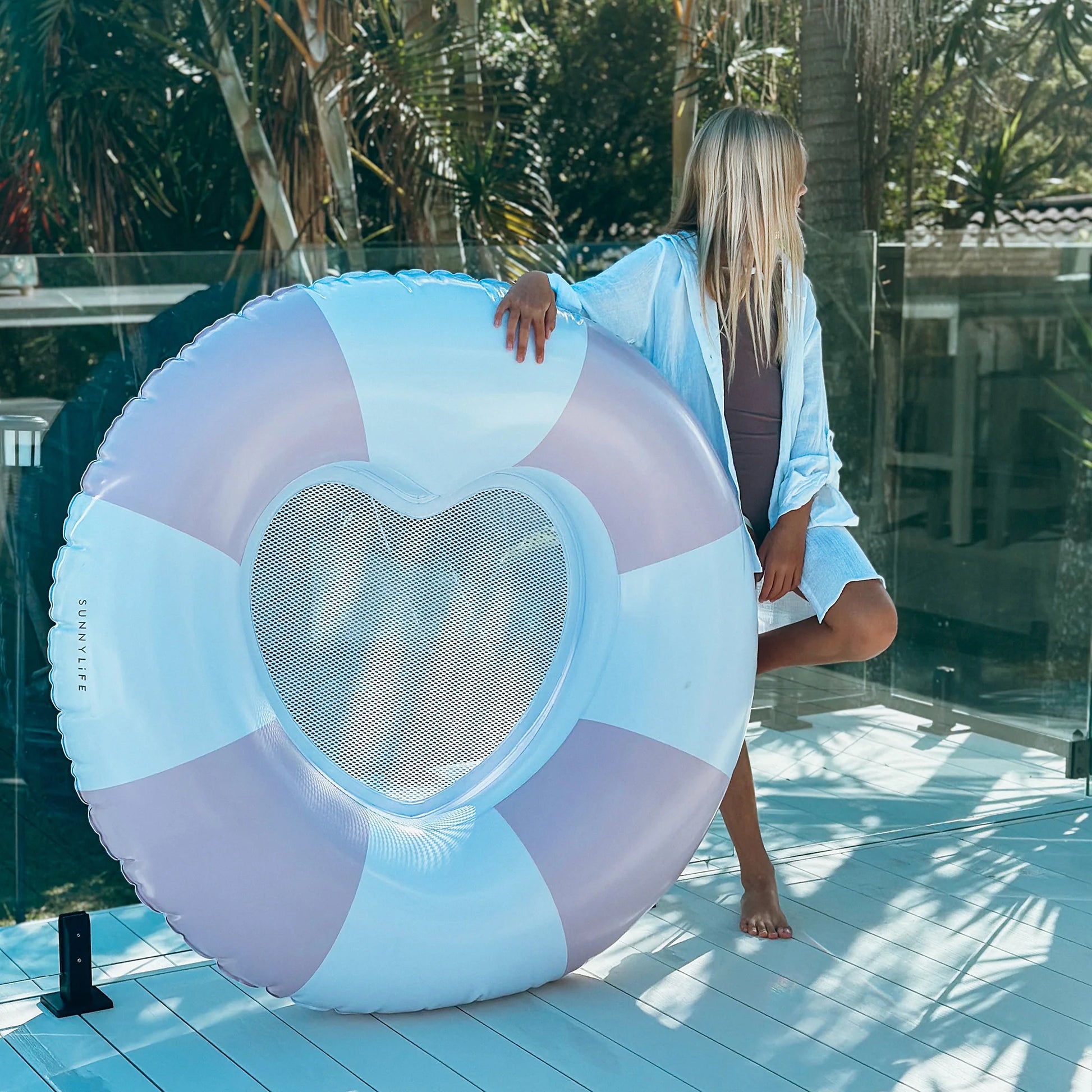 A girl in a light cover-up stands on a deck next to the Sunnylife Luxe Poolside Tube Float Pink Stripe, its heart-shaped center and pastel stripes capturing effortless Australian summer style. Palm trees sway in the background.