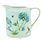 This light green ceramic creamer from the Aquazzura Secret Garden Collection by Aquazzura Casa features a garden motif adorned with vibrant illustrations of blue flowers, a peacock, a bee, and a dragonfly.