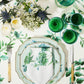 Set a charming table with Aquazzura Casa's Secret Garden Table Linen Collection: green and white floral-patterned plates, floral napkins, green glassware, cutlery, and flower arrangements atop matching linens for an elegant touch.
