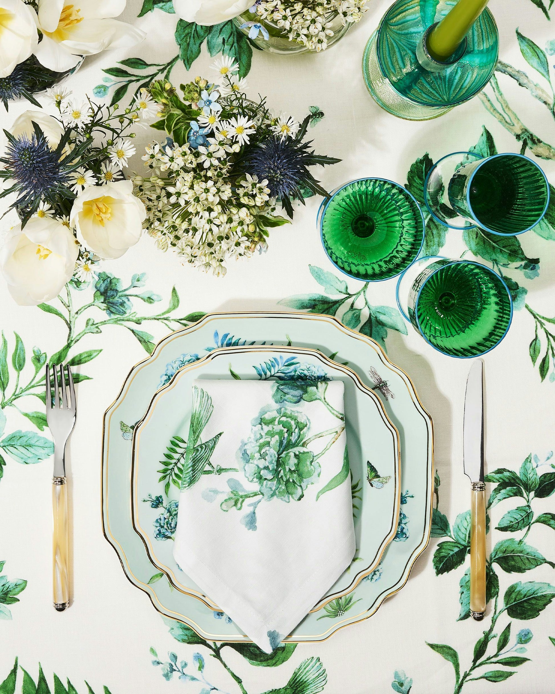 Set a charming table with Aquazzura Casa's Secret Garden Table Linen Collection: green and white floral-patterned plates, floral napkins, green glassware, cutlery, and flower arrangements atop matching linens for an elegant touch.