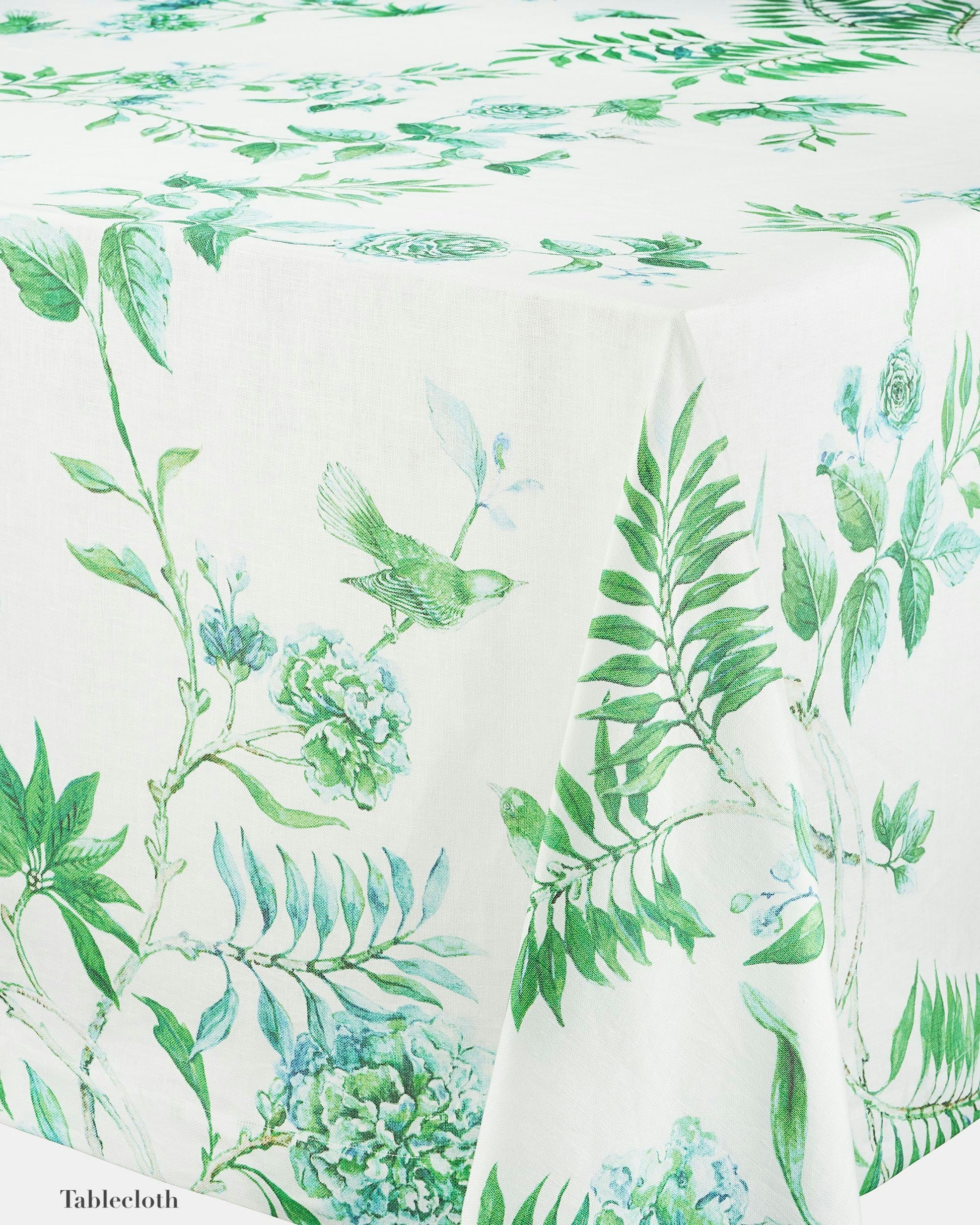 The Aquazzura Casa Secret Garden Table Linen Collection features a white tablecloth with green botanical and bird prints, ideal for a coordinated dining look when paired with matching placemats or napkins.