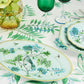 A table set with Aquazzura Casa's Secret Garden Table Linen Collection features floral plates, green glassware, matching placemats, a vase of white flowers, and a green-and-blue floral tablecloth.