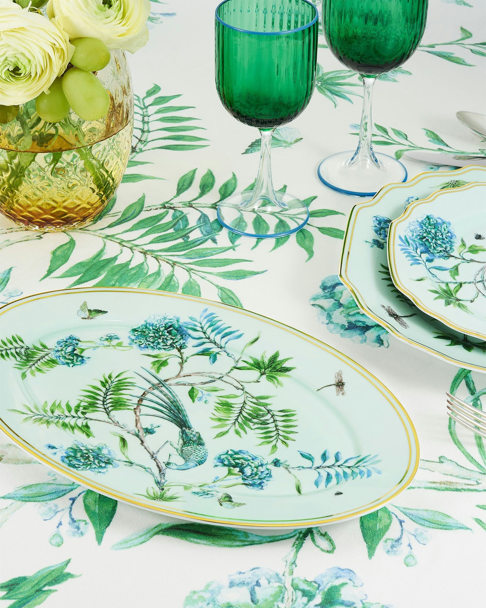 A table set with Aquazzura Casa's Secret Garden Table Linen Collection features floral plates, green glassware, matching placemats, a vase of white flowers, and a green-and-blue floral tablecloth.