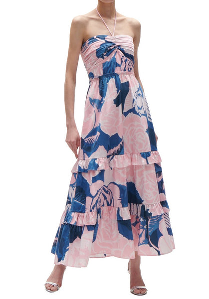 A woman stands wearing a halter, tiered maxi Figue June Dress with a pink and blue floral pattern, paired with white open-toe sandals. The background is plain and white.