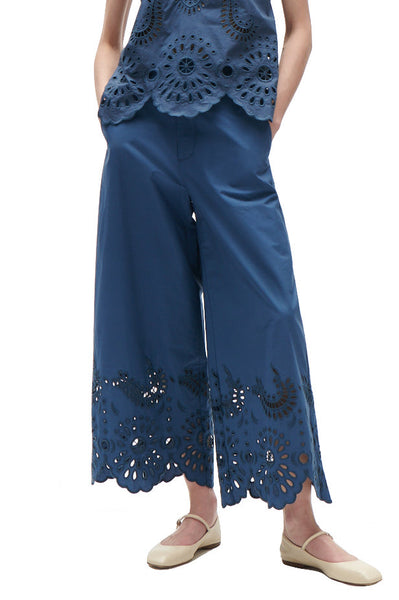 Woman wearing Figue Ramona Pant, a blue jumpsuit with cut-out patterns and embroidery detail, paired with beige flat shoes.