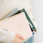 A person wears a cream sweater and blue jeans while holding several colorful spiral notebooks, with the top one—the Sugar Paper "Mom/Life" Spiral Notebook—featuring "MOM LIFE" elegantly stamped in gold. This chic notebook is perfect as an organized mom notebook or a guided planner for moms.