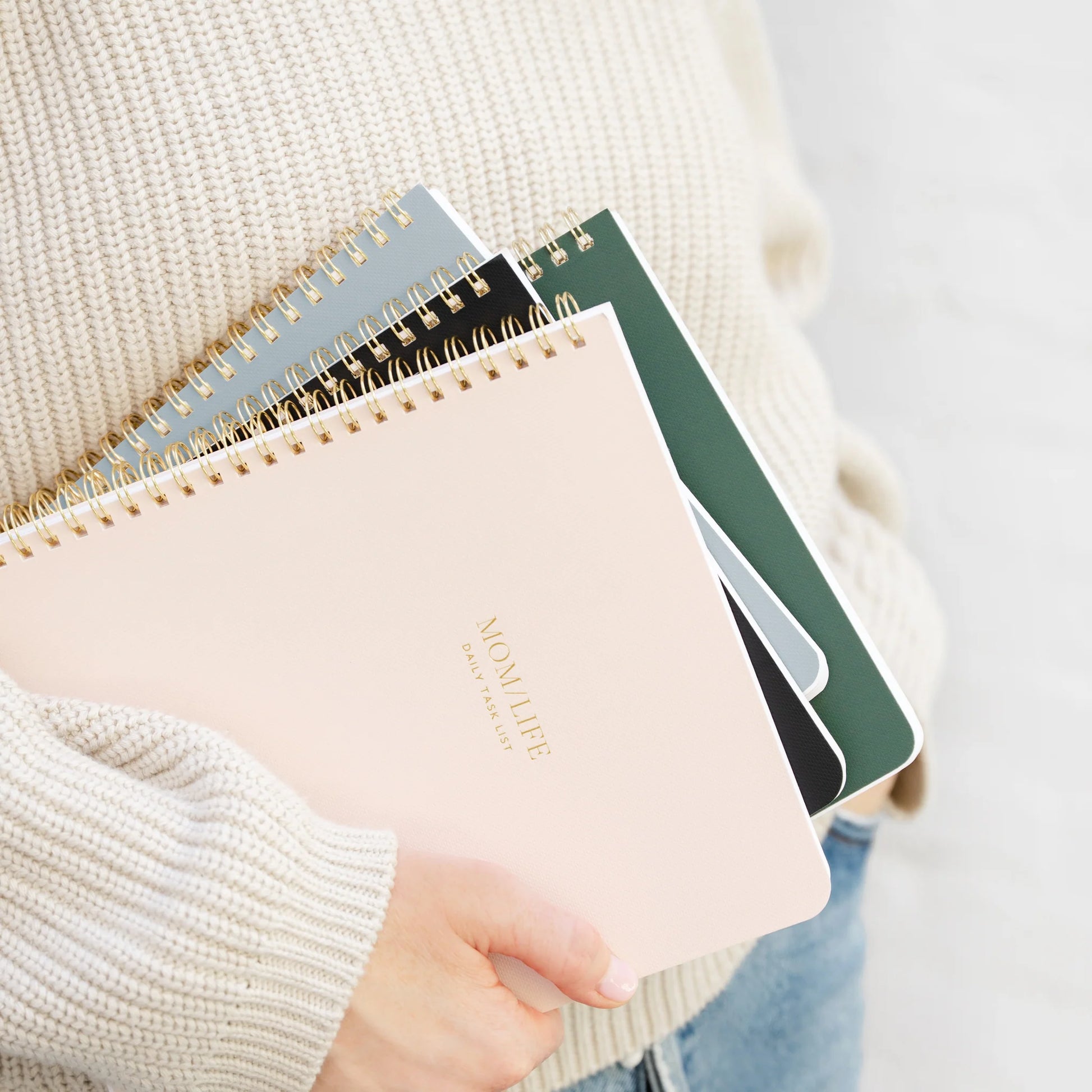 A person wears a cream sweater and blue jeans while holding several colorful spiral notebooks, with the top one—the Sugar Paper "Mom/Life" Spiral Notebook—featuring "MOM LIFE" elegantly stamped in gold. This chic notebook is perfect as an organized mom notebook or a guided planner for moms.