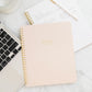 A Sugar Paper "Mom/Life Spiral Notebook" rests on a marble desk next to a laptop, a handwritten notepad, and a white cup with matching saucer—an ideal organizer for busy moms.