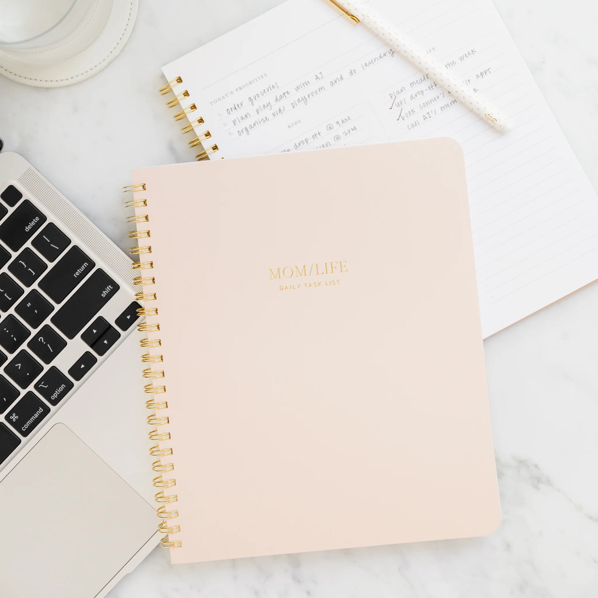 A Sugar Paper "Mom/Life Spiral Notebook" rests on a marble desk next to a laptop, a handwritten notepad, and a white cup with matching saucer—an ideal organizer for busy moms.