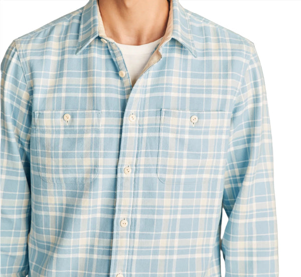 A person wears the Faherty The Surf Flannel shirt by Faherty, featuring a light blue plaid pattern, two chest pockets, and white buttons, made from organic cotton, over a white undershirt.