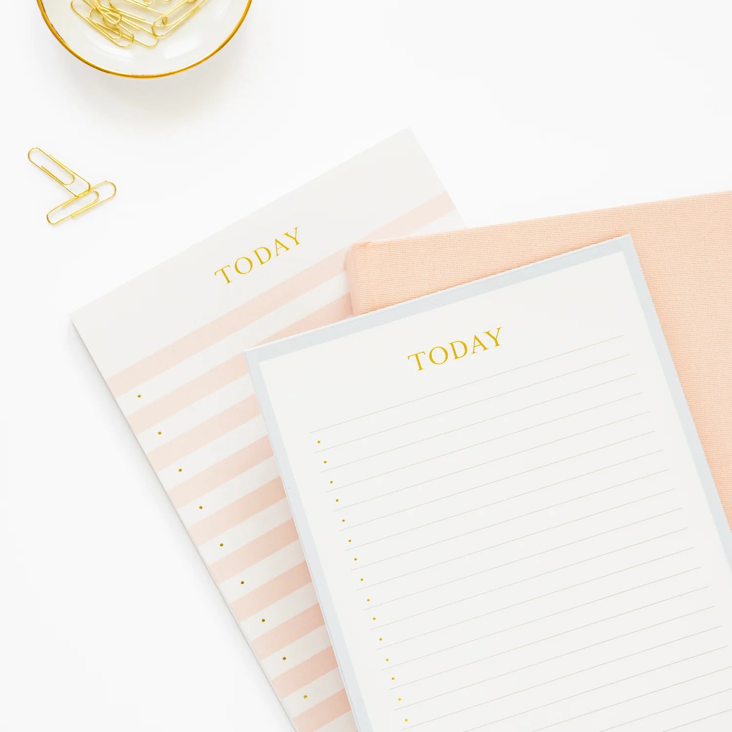 Two Sugar Paper Pink Striped Today Pads with gold lettering, a peach fabric item, and a dish with gold paper clips sit elegantly on a white surface—perfect for those seeking a chic daily notepad or blank canvas notepad to organize their day.