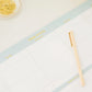 The Sugar Paper Desktop Family Planning Pad is a large-format weekly planner with spacious blank sections, accompanied by a gold pen and a dish of gold paper clips in the corner. Ideal for organizing your schedule, this pad also includes space for a daily menu section to help you keep track of meals throughout the week.
