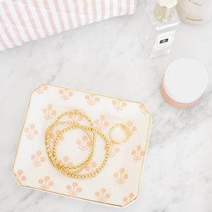The Sugar Paper Rose Floral Trinket Tray sits on marble holding gold jewelry; nearby are a striped towel, a glass perfume bottle, and a small cosmetic jar with gold accents.