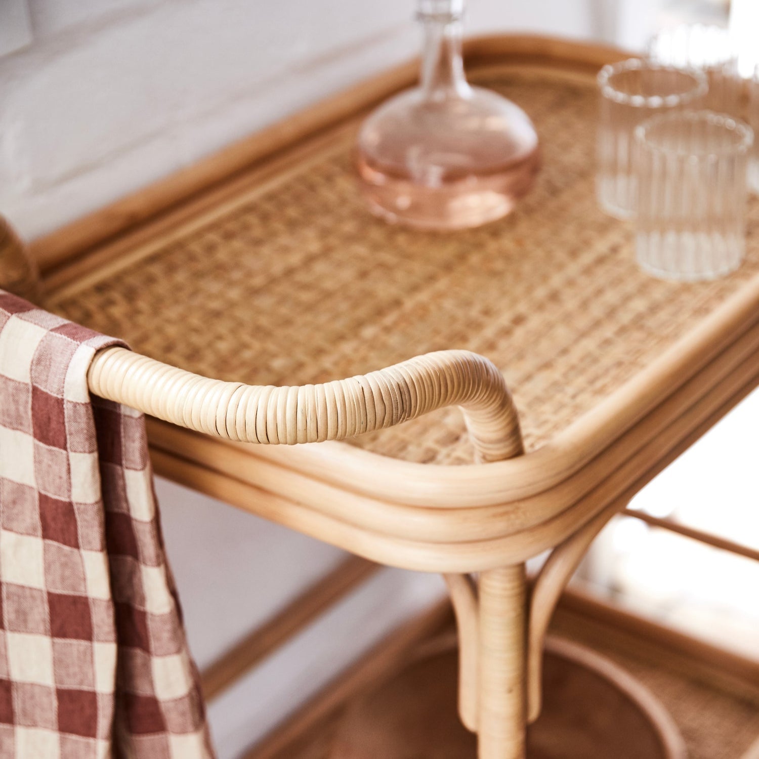 A close-up of the Selamat Flore Bistro Bar Cart in French café style, with a checkered cloth draped over the side, holding a glass decanter filled with pink liquid and two clear glasses.