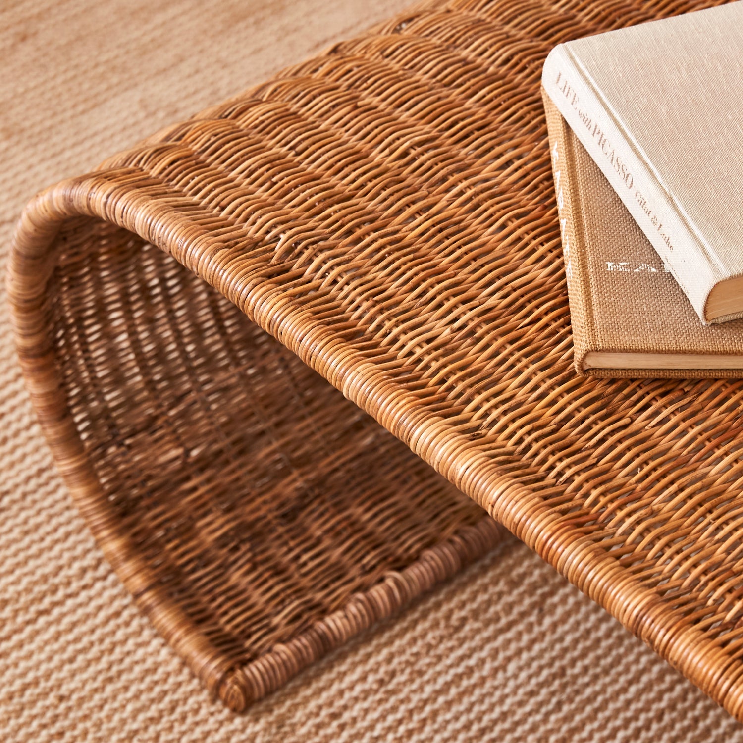 The Selamat Healdsburg Wicker Coffee Table, featuring a curved handwoven design, displays two closed books on top and sits on a woven rug with a diagonal stripe pattern.