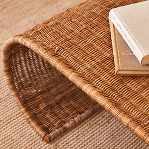 The Selamat Healdsburg Wicker Coffee Table, featuring a curved handwoven design, displays two closed books on top and sits on a woven rug with a diagonal stripe pattern.