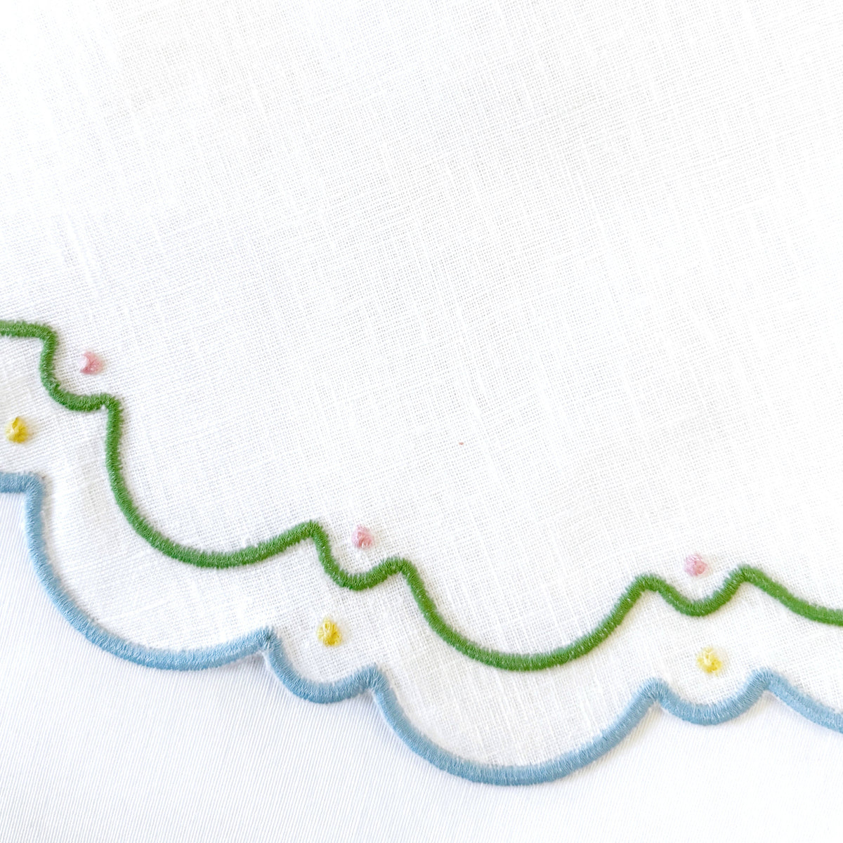 Sugus Table Linen Collection white napkins with scalloped edges embroidered in green and blue, accented with small pink and yellow stitched dots.