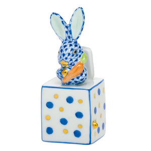 The Herend Jack in the Box Bunny is a hand-painted ceramic figurine featuring a blue patterned rabbit with an orange carrot, seated on a white cube with blue and yellow dots—perfect as a decorative accent for any space.