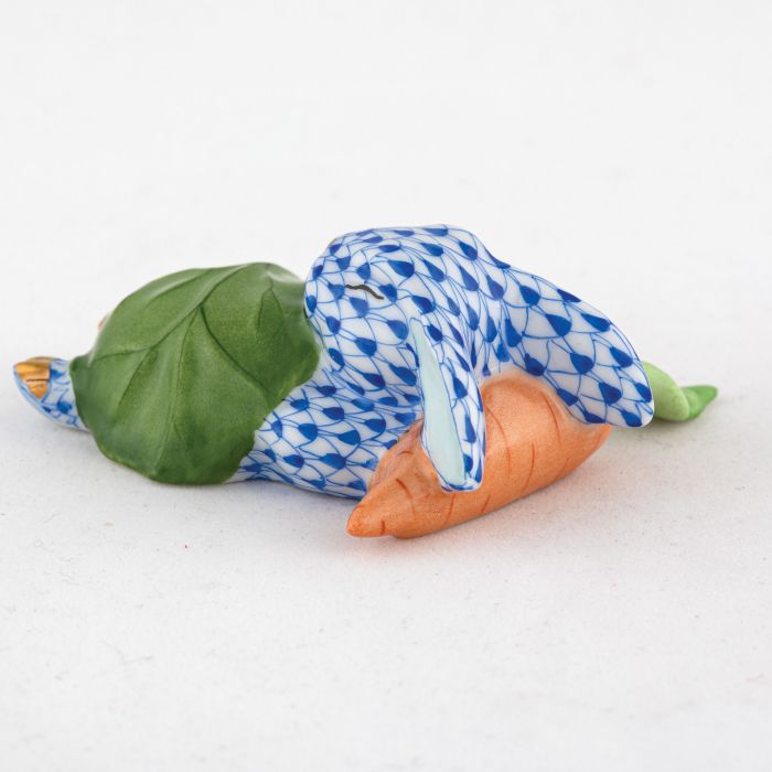 The Herend Slumber Bunny is a handcrafted Hungarian porcelain figurine featuring a blue fishnet motif rabbit lying on its side, holding a carrot, and detailed with a green leaf on its back, set against a plain white background.