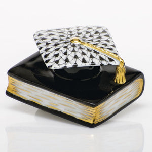 The Herend Graduation Cap is a collectible porcelain figurine featuring a black book with gold-edged pages and a graduation cap adorned with a gold tassel and black-and-white geometric pattern—a timeless decorative accent.