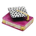 The Herend Graduation Cap is a decorative accent, featuring a pink and gold book topped with a black-and-white cap, adding timeless elegance and charm to any space.