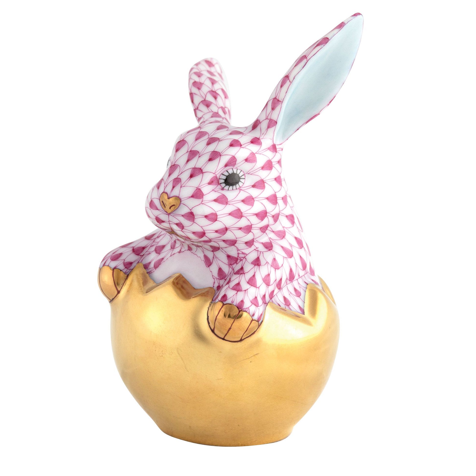 The Herend Hatching Bunny is a porcelain figurine depicting a pink-patterned rabbit emerging from a golden eggshell, with hand-painted details and gold accents on its paws and nose.