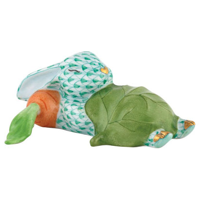 The Herend Slumber Bunny figurine, handcrafted in Hungary, depicts a patterned rabbit in the iconic fishnet motif, lying under a large green leaf with an orange carrot beside it.
