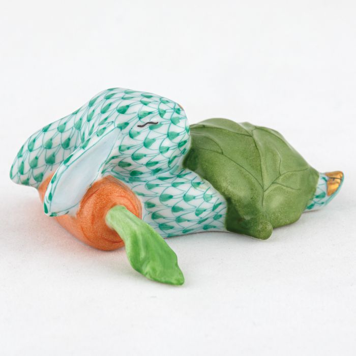 The Herend Slumber Bunny is a handcrafted porcelain figurine from Hungary, featuring a fishnet design. It rests on a green leaf, holding an orange carrot in its mouth, and is set against a white background.